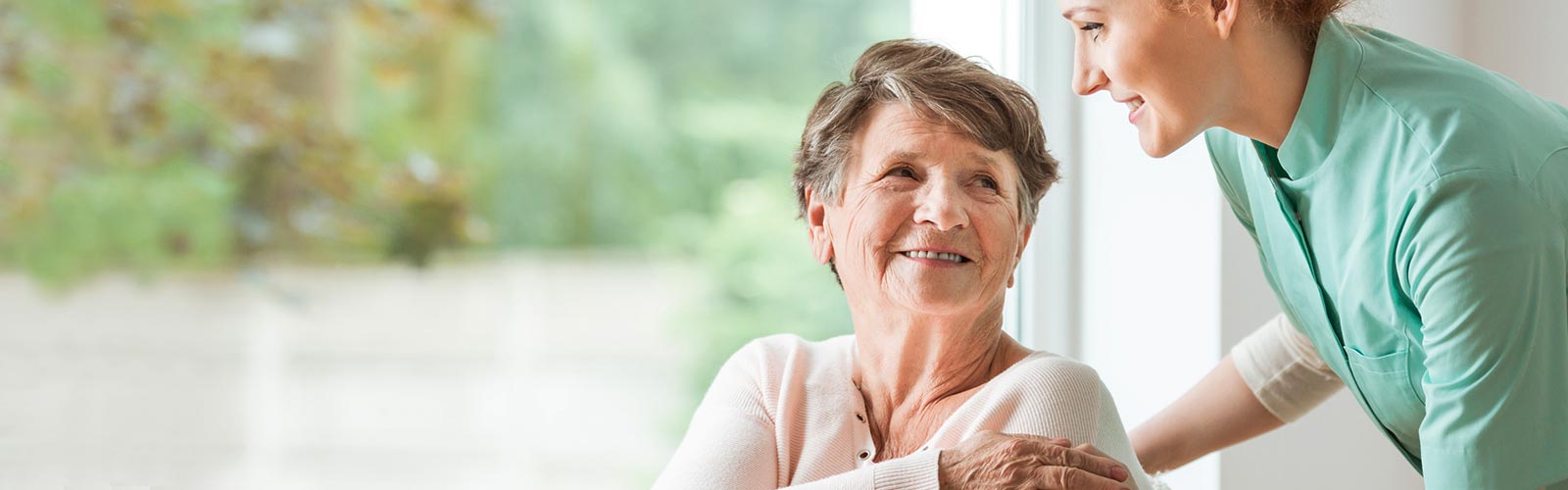 Pflegerin steht hinter der zu betreuenden Seniorin und lächelt sie an